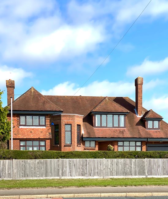 A large two-storey brick house with a pitched red tile roof, featuring multiple rectangular and bay windows with dark frames, surrounded by a well-maintained garden. The garden includes a neatly trimm