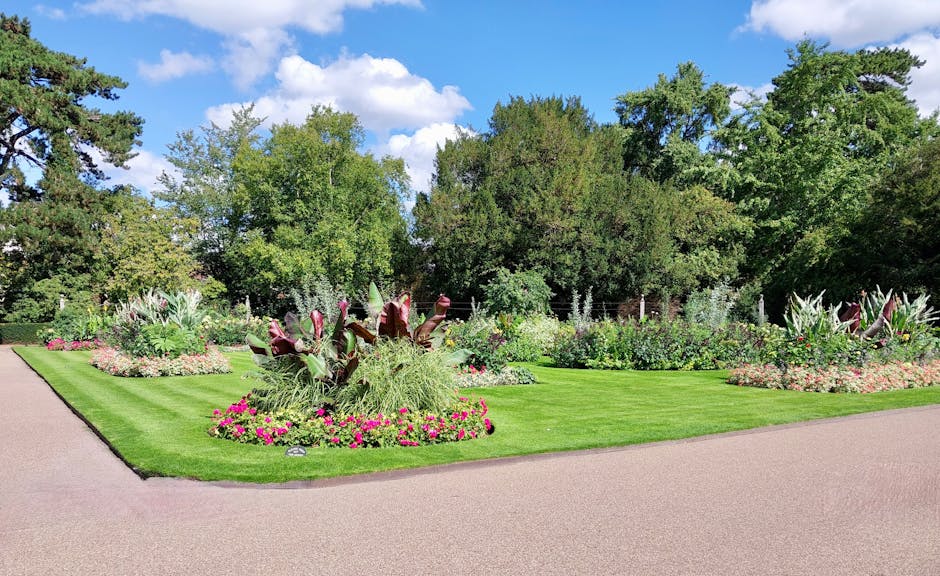 A wide view of a well-maintained garden featuring a lush, green lawn with neatly edged borders. In the center, there is a large flower bed populated with a variety of vibrant, flowering plants and orn